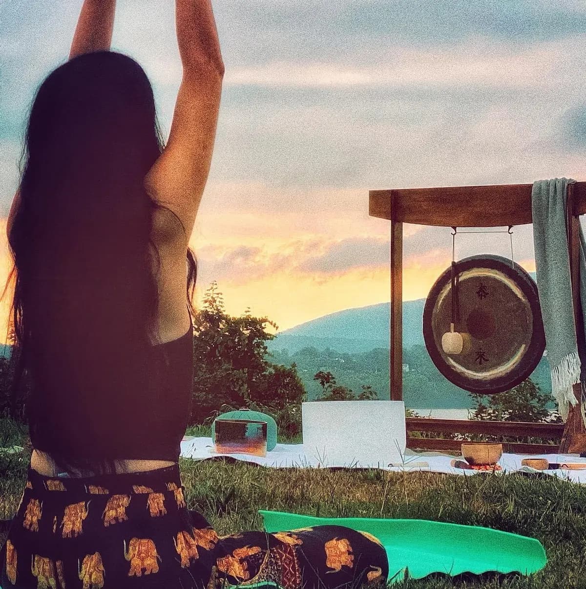 Una mujer con los brazos en alto frente a un gong en una colina al atardecer, con el valle del Hudson detrás.